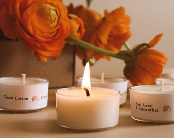May include: Four small, white tea light candles with labels. The candles are lit and the flames are visible. The labels read "Clean Cotton", "Clean", "Earl Grey & Cucumber", and "Earl Grey & Cucumber".