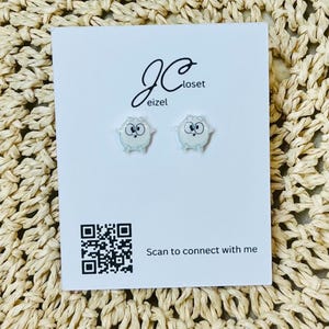 May include: A pair of owl-shaped stud earrings on a white card with the text "JC closet eizel". The card also features a QR code and the phrase "Scan to connect with me". The background is a woven, beige surface.