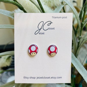 Puede incluir: Un par de pendientes de bot&oacute;n de champi&ntilde;&oacute;n rojo y blanco con postes de titanio. Los pendientes se muestran en una tarjeta blanca con el texto "Titanium post" y "Etsy shop: jeizelcloset.etsy.com".