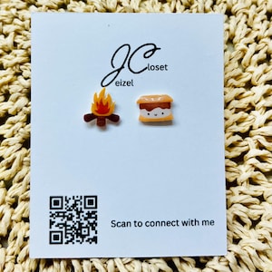 May include: A pair of novelty earrings on a white card. One earring is a campfire with orange and yellow flames, the other is a s'more. The card has the text "JC Closet eizel" and "Scan to connect with me".