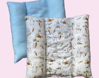 May include: A rectangular pillow featuring a white fabric side with a repeating pattern of line-drawn rabbits holding orange carrots with green tops, and a solid light blue fabric side. The pillow is constructed with three vertical sections.