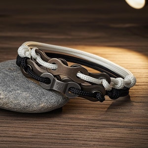 Könnte beinhalten: Drei Armbänder mit Fahrradkettendesign. Eines hat ein weißes Seil, ein anderes ein schwarzes Seil und das dritte ein schwarz-weißes Seil. Die Armbänder liegen auf einem grauen Stein, mit einem hölzernen Hintergrund.
