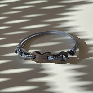 Puede incluir: Una pulsera gris con un dise&ntilde;o de cadena de bicicleta, con las letras "KMC" en el cierre. La pulsera est&aacute; hecha de un material gris similar a una cuerda y eslabones de cadena de metal. Un accesorio elegante.