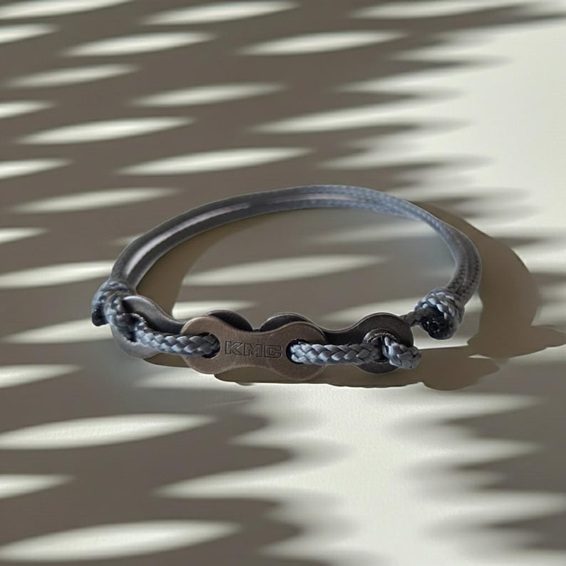 Puede incluir: Una pulsera gris con un dise&ntilde;o de cadena de bicicleta, con las letras "KMC" en el cierre. La pulsera est&aacute; hecha de un material gris similar a una cuerda y eslabones de cadena de metal. Un accesorio elegante.