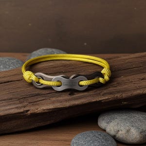 Puede incluir: Una pulsera con un dise&ntilde;o de cadena de bicicleta y un cord&oacute;n amarillo. Los eslabones de la cadena met&aacute;lica son de color gris oscuro y el cord&oacute;n es de color amarillo brillante. La pulsera se muestra sobre un trozo de madera.