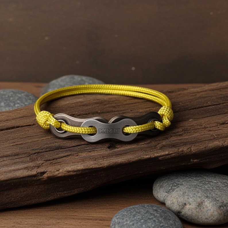 Puede incluir: Una pulsera con un dise&ntilde;o de cadena de bicicleta y un cord&oacute;n amarillo. Los eslabones de la cadena met&aacute;lica son de color gris oscuro y el cord&oacute;n es de color amarillo brillante. La pulsera se muestra sobre un trozo de madera.