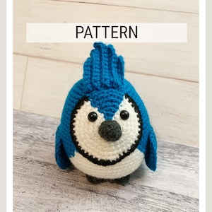 以下が含まれることがあります： 白いお腹、黒いアクセント、灰色のくちばしを持つ手作りのかぎ針編みの青い鳥。鳥の上には「PATTERN」の文字が表示されています。鳥は明るい灰色の木製の表面に座っています。
