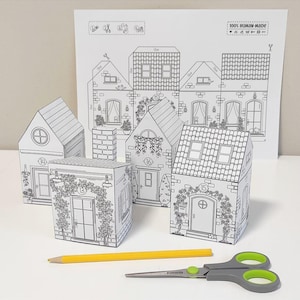 Puede incluir: Un kit de manualidades de papel en blanco y negro con diseños de casas en miniatura. El kit incluye casas ensambladas, una hoja impresa con plantillas de casas, un lápiz amarillo y tijeras. El texto "100% HUMAN MADE" es visible.