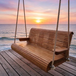 Puede incluir: Un banco columpio de madera con reposabrazos cuelga de gruesas cuerdas en un muelle de madera. El banco mira al mar al atardecer, con un cielo colorido de rosas, naranjas y amarillos. El banco es de madera marrón claro.