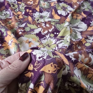 Op de afbeelding: Close-up van een stof met een bloemenpatroon. De stof heeft een dieppaarse achtergrond met een herhalend patroon van bloemen in tinten wit, groen, oranje en bruin. De stof is zacht en vloeiend.