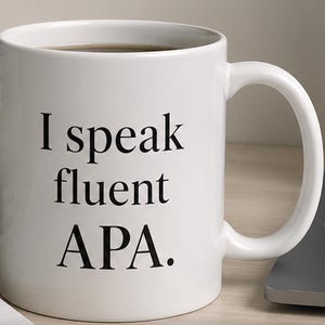Peut inclure: Mug en céramique blanche avec le texte "I speak fluent APA" en noir. La tasse a une anse incurvée et est remplie d'un liquide foncé, probablement du café ou du thé. La tasse est sur une surface claire.