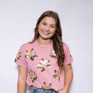 May include: A pink t-shirt with a floral sequin design. The short-sleeved shirt is paired with light blue jeans. The model is smiling and has long brown hair.