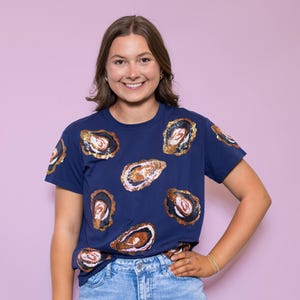 May include: Navy blue t-shirt featuring a pattern of oyster shell designs with gold, white, and black accents. The short-sleeved shirt is made of a soft material and is paired with blue jeans. The shirt is a unique fashion statement.