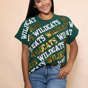 Könnte beinhalten: Grünes T-Shirt mit dem Wort "WILDCATS" in weißer und goldener Schrift. Das Shirt wird mit einer blauen Jeans kombiniert. Das Model lächelt.