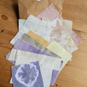 May include: A brown paper envelope containing a selection of fabric swatches in shades of purple, pink, yellow, and beige. The swatches are layered and overlapping, displaying a variety of hues and textures.