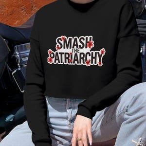 Może przedstawiać: Czarna bluza z długim rękawem z białym napisem "SMASH THE PATRIARCHY" otoczonym małymi czerwonymi kwiatowymi akcentami na klatce piersiowej. Odzież ma okrągły dekolt i skrócony dół, zestawiona z jasnoniebieskimi dżinsami.