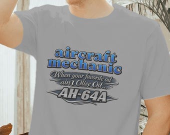 May include: A grey short-sleeved t-shirt featuring the blue and white text "aircraft mechanic" followed by the script text "When your favourite oil ain't Olive Oil..." and the bold text "AH-64A" centred on the chest area of the garment.