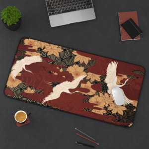May include: A rectangular desk mat featuring a Japanese-inspired design. The mat showcases a maroon background with white cranes in flight, complemented by gold and black floral and wave patterns. A white computer mouse rests on the mat.