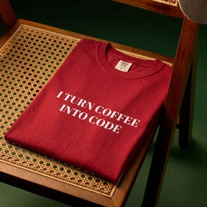 May include: A dark red short-sleeved t-shirt featuring the white printed text I TURN COFFEE INTO CODE on the front. The shirt has a crew neckline and a visible interior brand label at the collar, displayed folded on a wooden chair with cane seating.