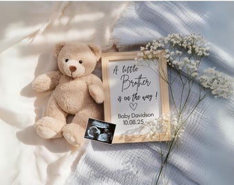 May include: A wooden sign with the text "A little brother is on the way!" with a heart and the date "10.08.25". The sign is surrounded by white flowers and a teddy bear.