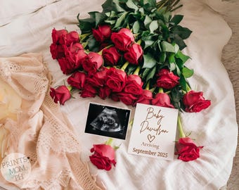 May include: A white card with the text "Baby Davidson Arriving September 2023" in gold lettering. A heart symbol is between the names and a black and white ultrasound image is in the background. The card is surrounded by red roses.