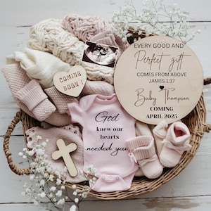 Puede incluir: Una cesta de mimbre llena de ropa de bebé, una cruz de madera, una manta blanca y un body rosa con el texto "Dios sabía que nuestros corazones te necesitaban". Hay dos letreros de madera con el texto "Todo buen y perfecto don viene de arriba Santiago 1:17 Bebé Thompson Llegada Abril 2025" y "¡Próximamente!"