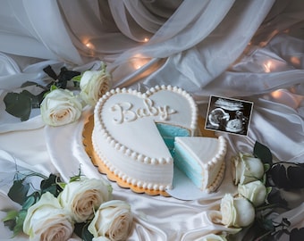 May include: A heart-shaped cake decorated with white frosting and the word "Baby" in cursive. The cake is cut open to reveal a blue layer inside. An ultrasound image is visible in the background.