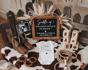 May include: A rustic baby announcement with a chalkboard sign that reads "Saddle Up! Our Grand Finale is on the Way! Our Herd is Complete September 2025". There is a white onesie with the text "Baby Brown" and a heart. There are also cowboy boots, a rope, and wooden blocks with letters.