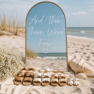 Peut inclure: Scène de plage avec un miroir doré portant l'inscription "And Then There Were Four". Plusieurs paires de sandales de différentes tailles sont disposées devant le miroir, avec un coquillage et des fleurs, sur le sable.