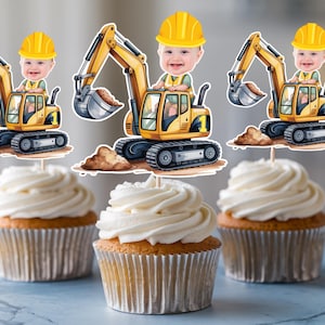 May include: Cupcakes topped with white frosting, each adorned with a construction-themed topper. The toppers feature a cartoon excavator with a baby wearing a yellow hard hat. The excavator is yellow and black.