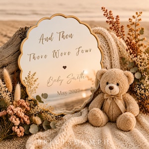 Puede incluir: Un espejo redondo de borde festoneado con marco dorado que presenta el texto And Then There Were Four, un corazón, Baby Smith y May 2027. Un oso de peluche marrón claro con un lazo de satén se sienta junto al espejo sobre una manta crema.