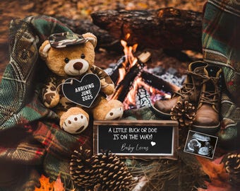 May include: A brown teddy bear wearing a camouflage hat holds a heart-shaped sign that says "Arriving June 2025". A wooden sign with the text "A little buck or doe is on the way! Baby Luens" is in the foreground. The image is set in a woodland scene with a campfire and pine cones.