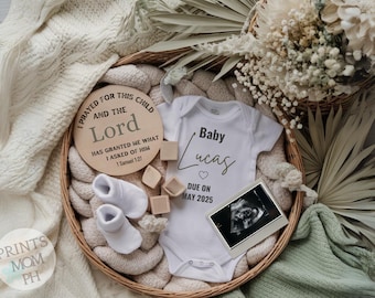 May include: A white onesie with the text "Baby Lucas" and a heart, along with the text "Due on May 2025". A wooden sign with the text "I prayed for this child and the Lord has granted me what I asked of Him 1 Samuel 1:27". A basket with a white blanket, white baby socks, and a framed ultrasound image.