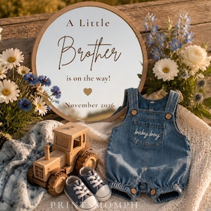 May include: A round wooden sign with the text A Little Brother is on the way! November 2026, a wooden toy tractor, a pair of small blue sneakers, and a denim romper with the words baby boy embroidered on the chest pocket, surrounded by flowers.