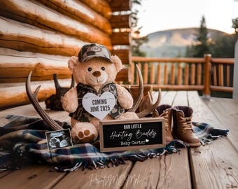 May include: A teddy bear wearing a camouflage hat and vest holds a sign that says "Coming June 2025". A wooden sign says "Our Little Hunting Buddy Baby Carter". The bear is sitting on a plaid blanket with antlers and boots.