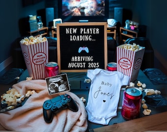Op de afbeelding: Een zwangerschapsaankondiging met een gamingthema, met popcorn, frisdrank en een babyromper. Een letterbord leest "NEW PLAYER LOADING... ARRIVING AUGUST 2025." Een echo en een gamecontroller zijn ook aanwezig.