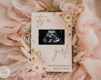 May include: A pink and white gender reveal announcement card with a sonogram image, the text "it's a girl!" and "Arriving Due on March 2025". The card is decorated with white daisies and dried flowers.