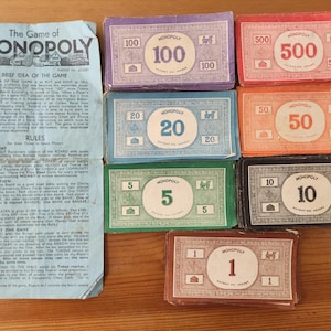 May include: A vintage Monopoly board game set with various denominations of play money and game instructions. The money is in purple, red, blue, orange, green, and black, with values from 1 to 500. The game instructions are on a light blue sheet.