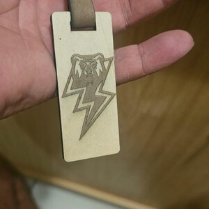 May include: Wooden keychain with a brown leather strap and silver keyring. The rectangular wooden tag features a laser-etched design of a bear's head inside a lightning bolt. The leather strap is secured with a metal snap.