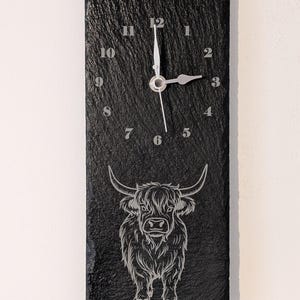May include: Black slate clock with a white clock face and silver hands. The clock features a laser-etched design of a Highland cow.