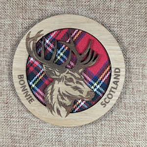 Könnte beinhalten: Ein runder Untersetzer aus Holz mit einem rot-schwarz-weißen Karomuster und einem Ausschnitt eines Hirschkopfes. Der Text "Bonnie Scotland" ist um den Rand des Untersetzers geschrieben.
