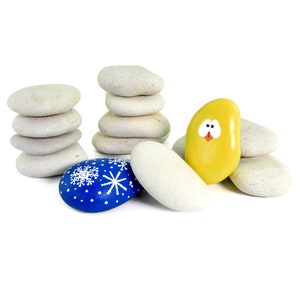 May include: A collection of painted rocks, including a yellow rock with a painted face, a blue rock with white snowflakes, and several white rocks stacked in a pile.