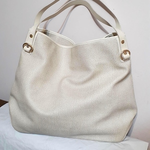 May include: Cream-colored tote bag with a textured surface and two shoulder straps. The bag features gold-colored metal accents at the strap attachments. The bag is a neutral color and has a spacious design.