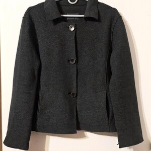 Op de afbeelding: Een donkergrijze blazer met een klassieke kraag en drie knopen. De jas hangt aan een zilveren hanger tegen een lichte achtergrond. De blazer heeft een getailleerde pasvorm en lijkt gemaakt van een wolmix.