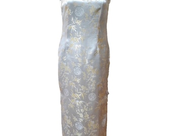 May include: A sleeveless, floor-length, ivory-colored dress with a mandarin collar. The dress features a subtle floral and bamboo leaf pattern in gold. The dress is displayed on a black mannequin.