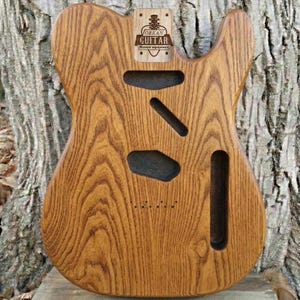 Può includere: Un corpo per chitarra elettrica in legno non rifinito con un evidente motivo a venature naturali. Il corpo presenta cavità pretagliate per i pickup e l'elettronica, e una placca rettangolare con il testo ORKAN GUITAR Musical equipment.