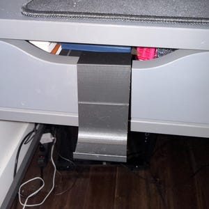 May include: A black, 3D-printed desk drawer organiser is attached to a grey desk drawer. The organiser is rectangular and has a curved bottom. The drawer is partially open, revealing books and other items inside.