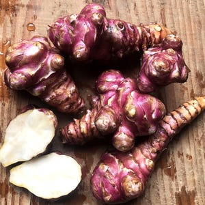 Op de afbeelding: Een bovenaanzicht van verschillende aardperen, met hun kenmerkende knobbelige, rood-paarse schil. Twee gesneden stukken onthullen een wit interieur. De groenten zijn gerangschikt op een verweerd houten oppervlak, wat hun unieke textuur en kleur benadrukt.