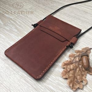 May include: A brown leather phone case with a black cord for wearing around the neck. The case has a flap closure and is stitched around the edges. The brand name "SG Leather" is embossed on the flap. An acorn and a dried leaf are next to the case.