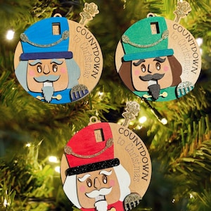 Può includere: Tre decorazioni natalizie in legno con disegni di schiaccianoci. Ogni ornamento ha un cappello di colore diverso: blu, verde e rosso. Gli ornamenti hanno un calendario alla rovescia e le parole "Countdown to Christmas".